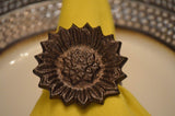 Dark bronze sunflower-shaped napkin ring holding a yellow cloth napkin, placed on a white plate with a decorative silver rim.