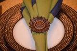 Brown sunflower-shaped napkin ring holding folded green and blue napkin on a round white plate with a textured dark brown rim.