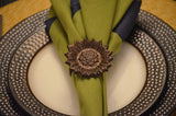 Antique bronze sunflower napkin ring holding an olive green and black cloth napkin, placed on a white plate with a textured black outer rim.