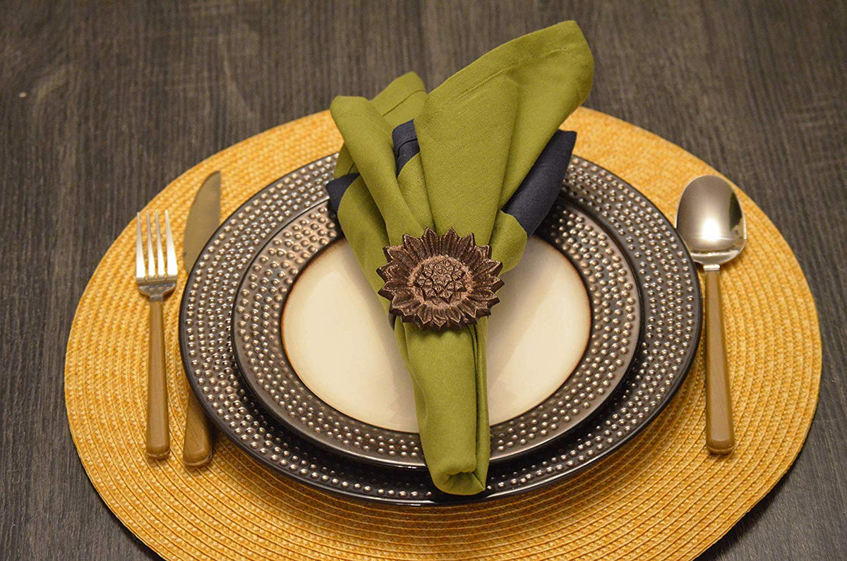 Table setting with cream and dark patterned plates on a yellow woven placemat, featuring green and dark napkins secured by a brown sunflower-shaped napkin ring.