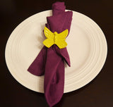 Yellow butterfly-shaped napkin ring on a folded purple cloth napkin, placed on a white round plate with concentric circle patterns, against a dark background.
