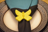 Yellow butterfly-shaped napkin ring holding a folded dark green napkin on a beige plate with a textured dark brown rim.