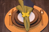 Yellow butterfly-shaped napkin ring holding a two-tone green and blue cloth napkin on white and brown dotted plates with gold cutlery and a woven round placemat.