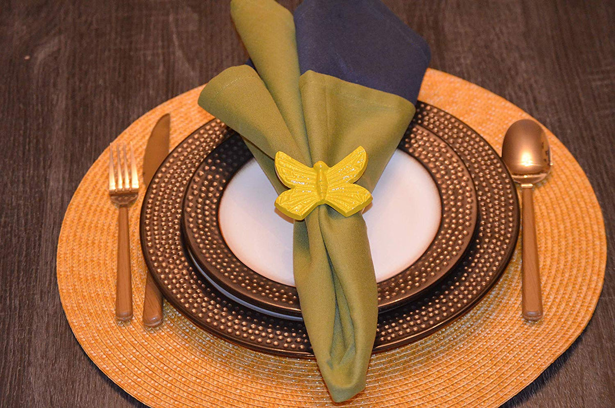 Yellow butterfly-shaped napkin ring holding a two-tone green and blue cloth napkin on white and brown dotted plates with gold cutlery and a woven round placemat.