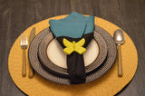 Table setting with blue and black cloth napkin held by a yellow butterfly-shaped napkin ring on stacked white and textured gray plates, with gold flatware on a woven placemat.