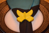 Yellow butterfly-shaped napkin ring holding a black cloth napkin on a white plate with a textured dark brown rim.