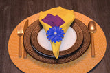 Yellow folded napkin with a purple fabric insert held by a blue flower napkin ring on stacked white and brown dotted plates, with gold-toned cutlery on an orange woven placemat.