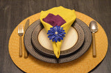 Yellow cloth napkin folded within a blue floral napkin ring with a white center, placed on stacked textured black and white plates, set on a round woven yellow placemat.