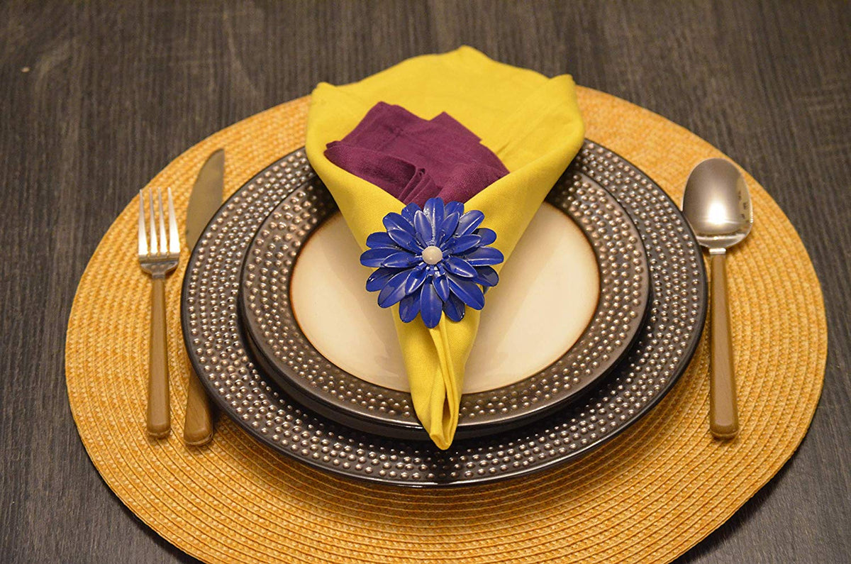 Yellow cloth napkin folded within a blue floral napkin ring with a white center, placed on stacked textured black and white plates, set on a round woven yellow placemat.