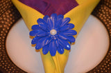 Bright blue flower-shaped napkin ring with detailed petals and a central pearl bead holding a yellow and purple folded napkin on a white plate with textured brown rim