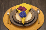 Table setting with a yellow napkin topped by a purple folded napkin secured by a blue floral napkin ring, on white and decorative black plates over a circular woven placemat.