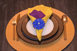 Round woven beige placemat with brown textured dinner and salad plates, white center plate, yellow and purple folded napkins, and blue floral napkin ring with a white center bead.