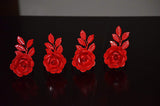 Set of four red napkin rings shaped like roses with detailed petals and three textured leaves on each, arranged in a row on a dark reflective surface.