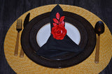 Black napkin folded on a white plate within a dark textured charger, accented with a red rose napkin ring, all on a round woven yellow placemat.
