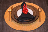 Table setting featuring a black napkin folded with a red rose napkin ring on a white plate with brown decorative plates, placed on a round woven tan placemat with gold flatware.