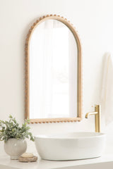 Arched wall mirror with a natural wood frame featuring rounded bead detailing, mounted above a white oval vessel sink with a brass faucet and potted greenery nearby.