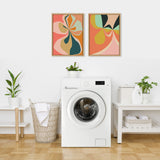 Modern laundry room with white front-load washer, two colorful abstract floral canvases in wood frames above, and green plants on white shelves and basket.