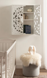 White carved wood wall cabinet with intricate floral patterns, partially open to reveal shelves holding toys and books, above a furry-topped gray stool in a nursery.