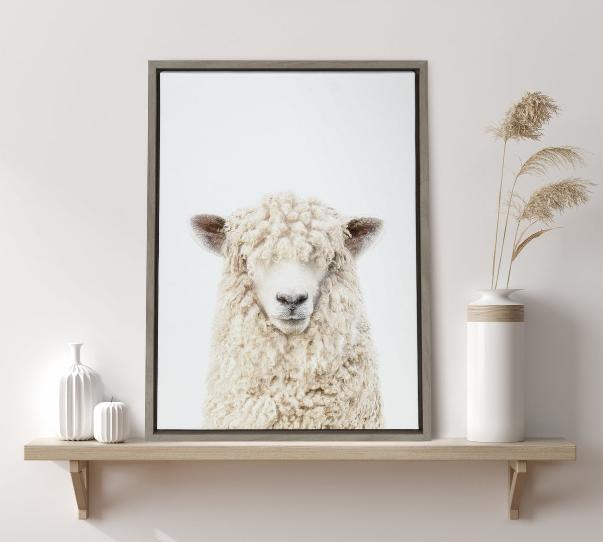 Framed canvas portrait of a white sheep with curly wool and bangs covering its eyes, displayed on a wooden shelf with neutral-toned vases and dried grasses.