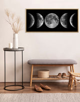 Framed black canvas displaying five realistic moon phases, hanging above a minimalist bench with wooden legs and grey cushion, next to a black metal side table with dried plants.