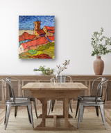 Colorful canvas print of a village with red roofs, a tower, and green hills under a blue, cloudy sky, hung above a wooden dining table with metal chairs.