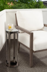 Tall cylindrical metal drink table with a dark bronze finish next to a cushioned outdoor chair with light cream fabric and wooden frame on a gray patio floor.