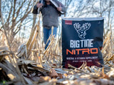 Black and orange Big Tine Nitro mineral and vitamin supplement bag standing upright on dry corn stalks with a blurred figure holding a crossbow in the background.