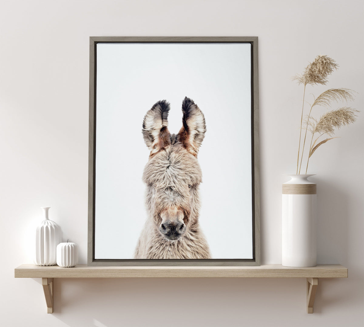 Framed canvas portrait of a burro with fluffy light gray and brown fur, tall ears with black tips, against a plain white background, displayed on a wooden shelf.