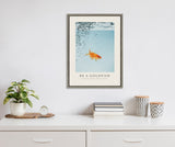 Framed art print showing a goldfish swimming upwards to clear blue water with scattered black dots, displayed above a white dresser with plant and books.