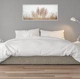 Minimalist bedroom with white bedding and pillows on a light gray upholstered bed frame, featuring a framed canvas of beige pampas grass against a pale background above the bed.