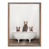 Three brown bear cubs sitting inside a white vintage clawfoot bathtub on a wooden floor, against a plain light background, with the center cub standing taller.