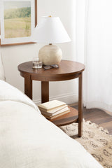 Round wooden side table with a warm brown finish, featuring a white ceramic lamp with a brass accent and a glass of water on top, plus books on the lower shelf.