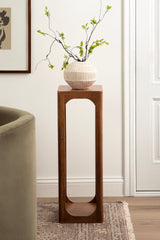 Tall, narrow Dahl Square Wood Tea Table with rounded rectangular cutout base holding a ribbed white ceramic vase with green leafy branches, placed on a beige rug near a green chair and framed wall art.