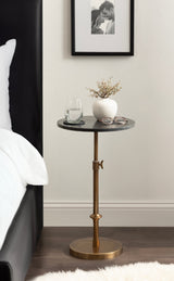 Small round black marble tabletop on an adjustable brass stand with a white ceramic vase holding twigs, a glass of water, and eyeglasses beside a black bed frame and white bedding.