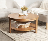 Round wooden coffee table with lower shelf holding a woven basket, placed on a patterned beige rug between two light gray upholstered chairs with white blankets.