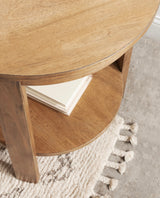 Close-up of a round wood side table with a natural finish, featuring a lower shelf holding stacked white books, standing on a textured beige rug with tassels.