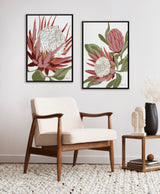 Two framed canvas artworks showing stylized King Protea flowers with red and white petals and green leaves, hung side by side above a mid-century modern wooden armchair.