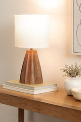 Table lamp with a white cylindrical shade and a scalloped, wood-toned base sitting atop stacked books on a wooden surface with a potted plant and light wall art nearby.