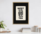Black and white framed artwork depicting a stylized playing card with a Queen holding a flower, captioned 'LUCKY YOU' above and below on a black matte background.
