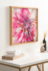 Bright pink and purple abstract floral artwork with dynamic swirling patterns, framed in light wood and hung on a white wall above a table with towels and vases.