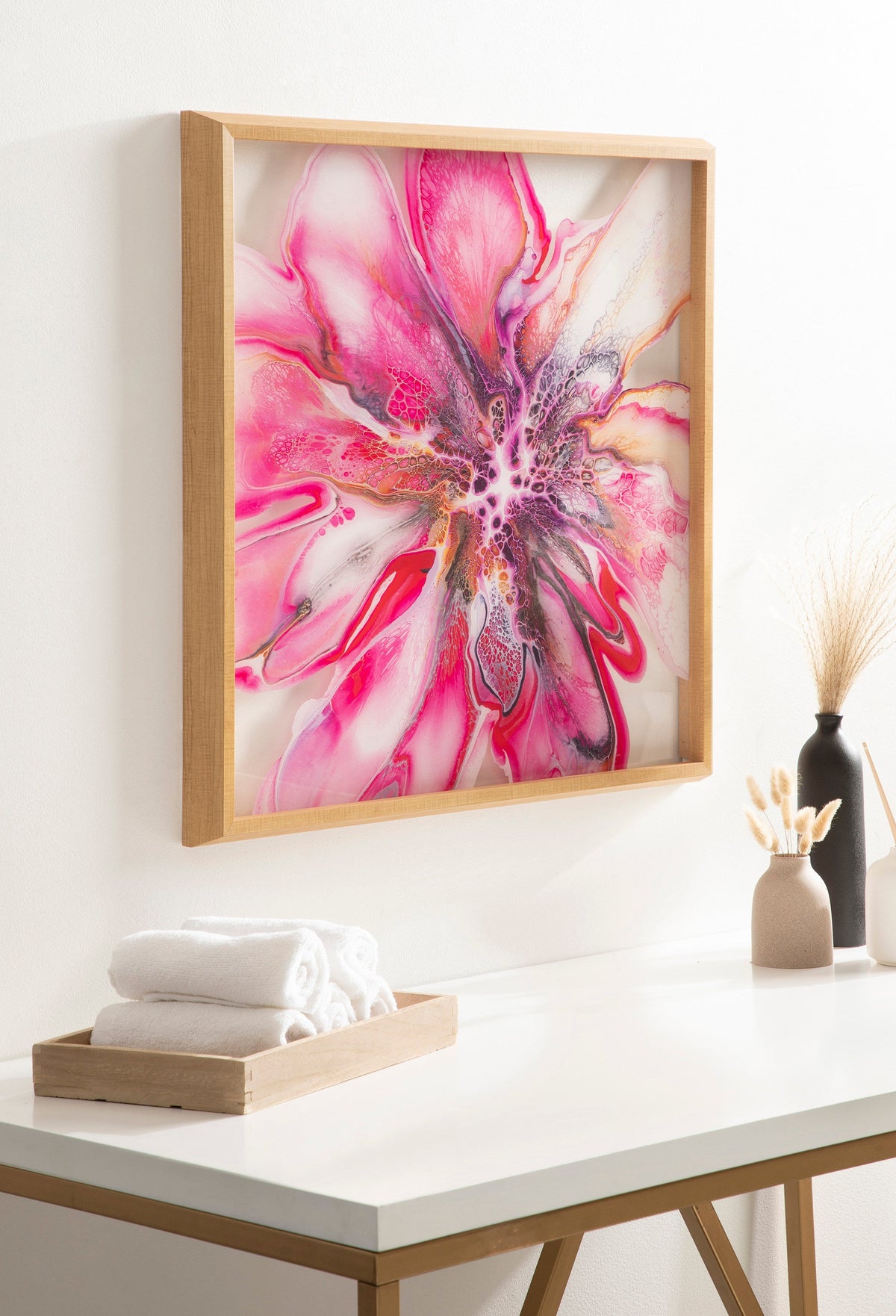 Bright pink and purple abstract floral artwork with dynamic swirling patterns, framed in light wood and hung on a white wall above a table with towels and vases.