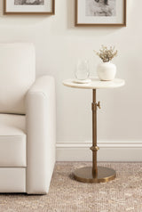 Round white marble table top on adjustable bronze metal stand beside a white upholstered sofa, with a glass of water and a small white vase with dried flowers on the table.