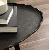 Close-up of a black textured oval tray with a wavy edge, holding two stacked black-covered books and a white ceramic cup on a wooden surface.