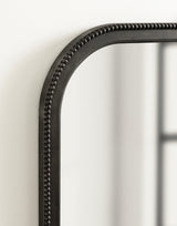 Close-up of a black arched wall mirror corner with a beaded trim detail on a white wall, reflecting a blurred window frame.