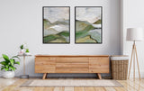 Two framed abstract canvases with soft green, beige, and gray hills and sky tones hang side by side above a light wood sideboard in a modern room.