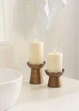 Two lit pillar candles on bronze metal holders with flared scalloped edges, displayed on a white bathroom countertop with patterned wallpaper.