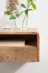 Close-up of a smooth-edged floating wooden side table with a natural grain, holding two clear glass vases containing green leafy branches against a white wall.
