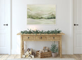 Muted green and beige winter landscape painting above rustic wood console table decorated with pine garland and candles, with logs and a small pine tree below.