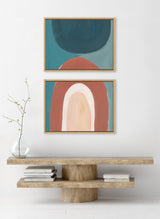 Two framed abstract canvas artworks above a minimalist wooden console with shelf; top piece has teal and dark gray shapes, bottom piece features layered arches in terracotta, cream, and teal.