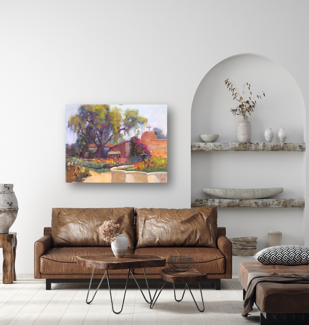 Colorful impressionist painting showing a garden path curving past a tree and building with a cross, displayed above a brown leather sofa in a bright room with curved niche shelves.